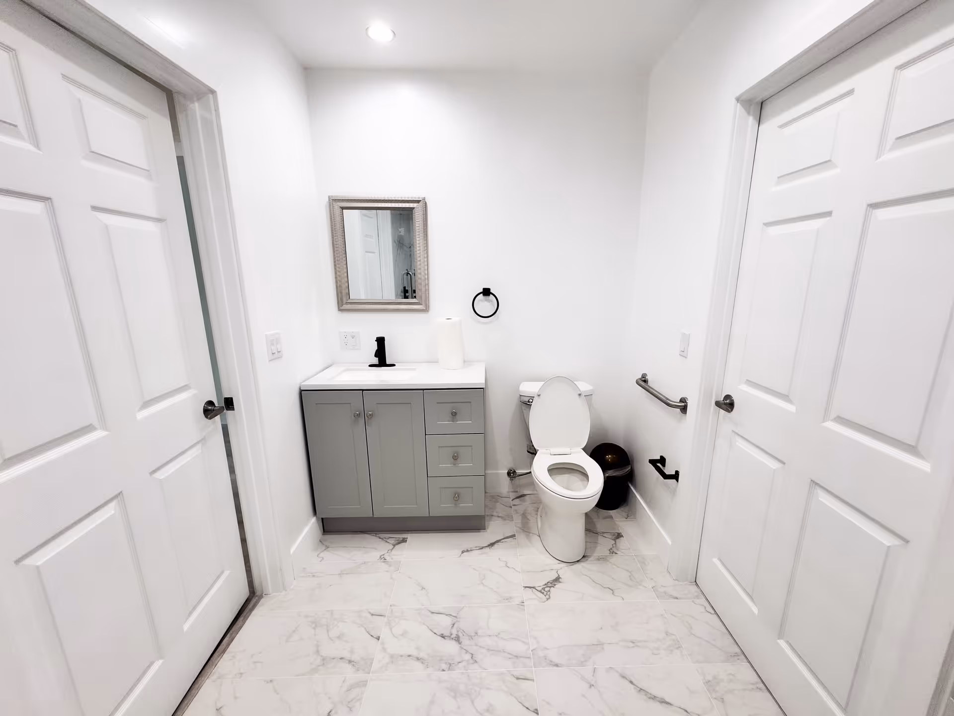 A clean, modern bathroom with white walls and marble tile flooring. It features a gray vanity with a white countertop and a black faucet, a small rectangular mirror above the sink, and a toilet with the lid open. There are two white paneled doors on either side of the bathroom, a black towel ring, a black toilet paper holder, and a metal grab bar on the wall next to the toilet.