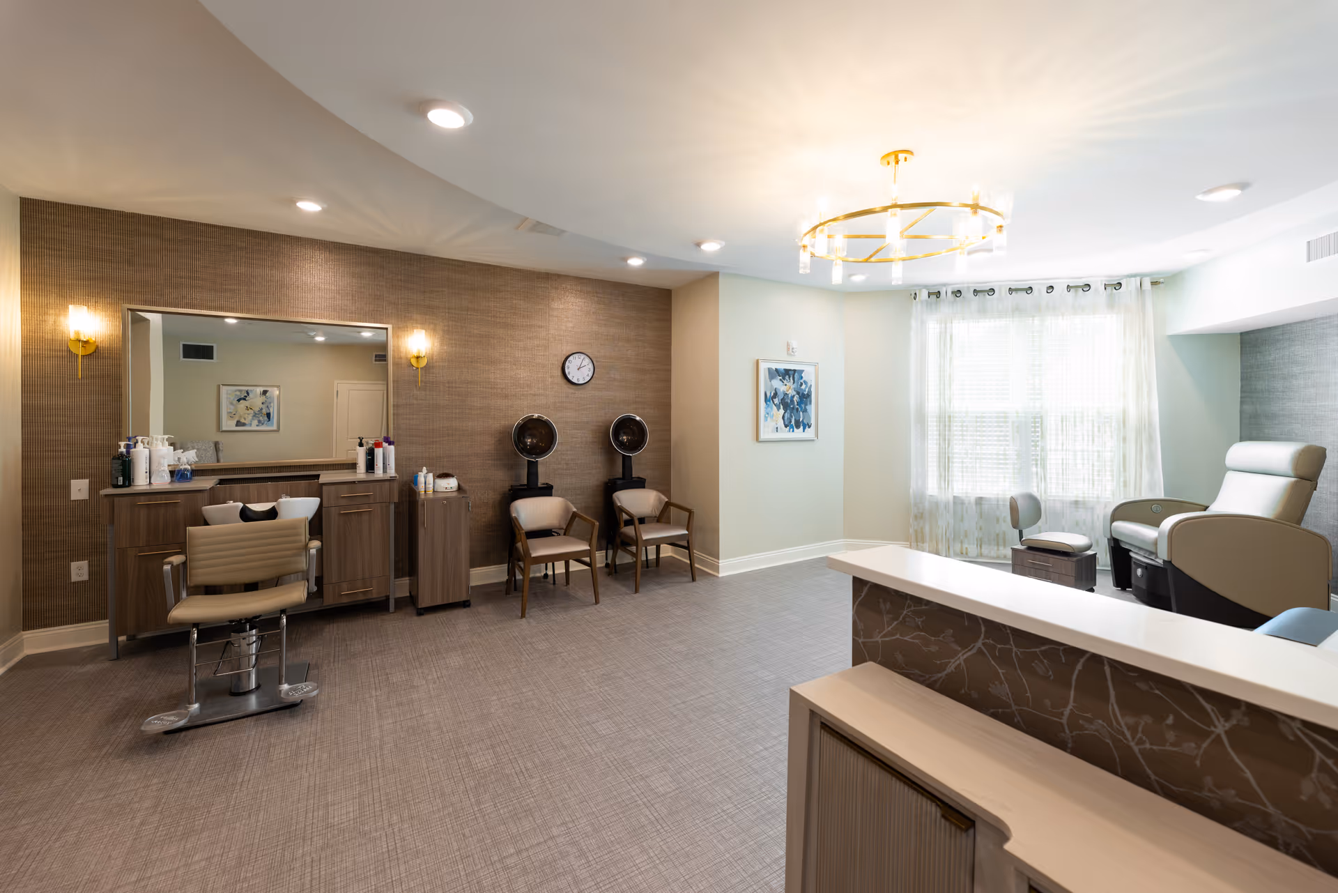 A spacious salon area in a senior living facility featuring a styling chair in front of a large mirror with hair care products on the counter, two hair drying chairs against a textured wall with a clock above, and a pedicure station with a comfortable chair near a window with sheer curtains. The room is well-lit with recessed lighting and a modern chandelier.