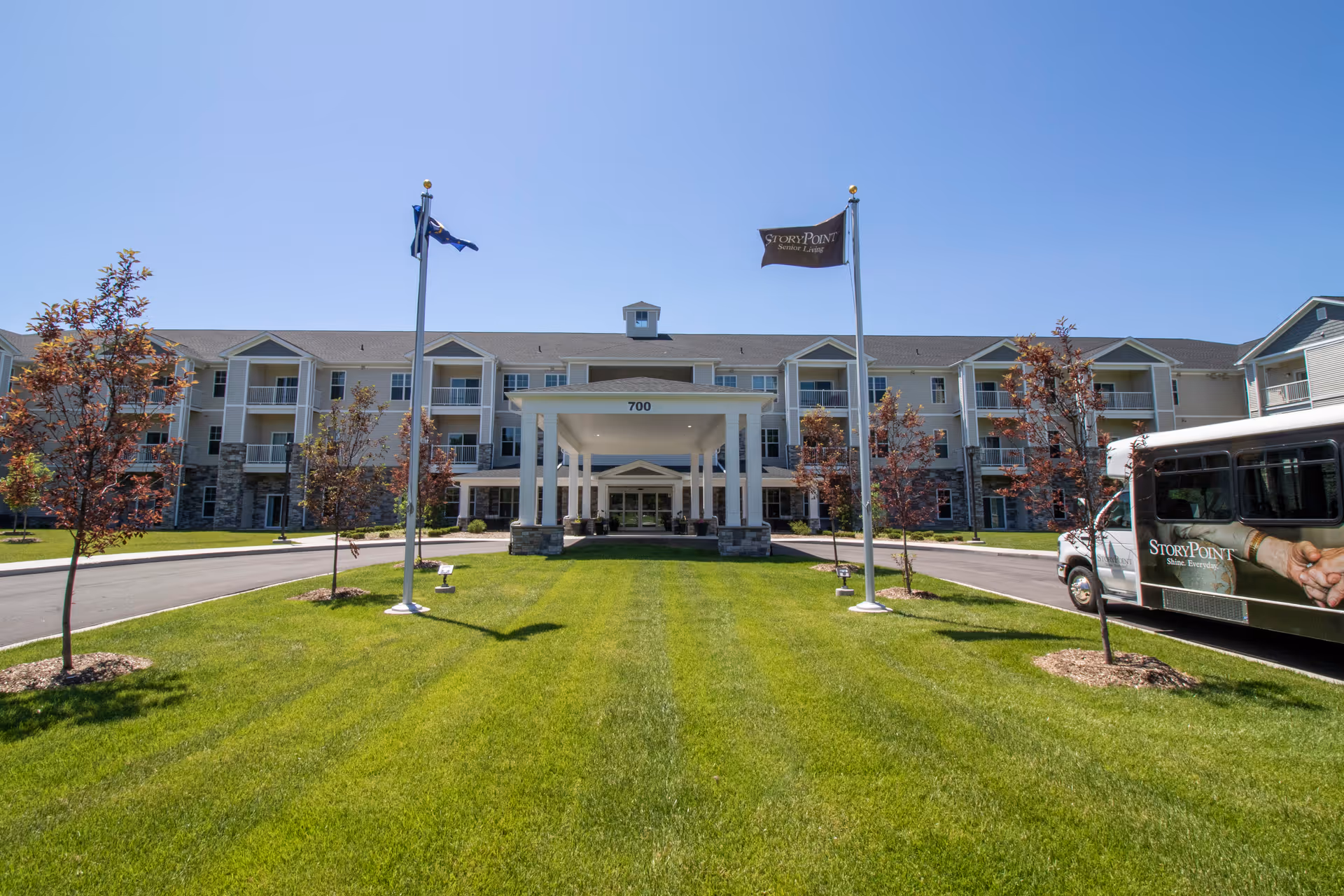 Front exterior of the StoryPoint Chesterton senior living building with a porte-cochere, flagpoles, manicured lawn, and a StoryPoint shuttle van.