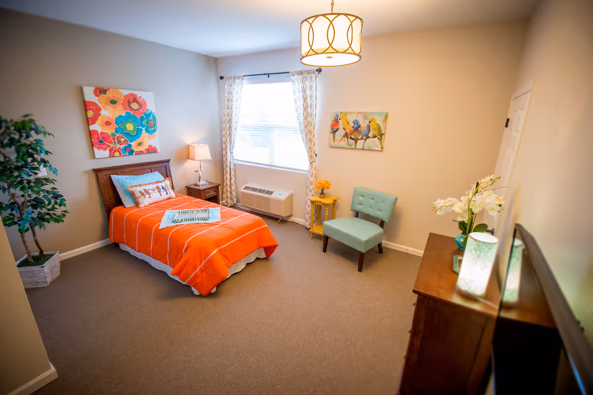 A cozy bedroom with a single bed covered in an orange blanket and colorful pillows. There is a nightstand with a lamp beside the bed, a large window with patterned curtains, a small turquoise chair next to a yellow side table with a flower vase, and a wooden dresser with decorative items including a lamp and flowers. The walls are decorated with bright floral and bird paintings, and there is a potted plant in the corner.