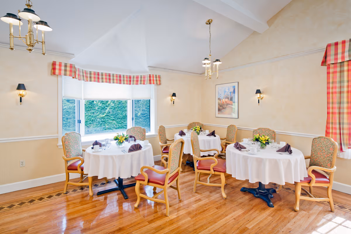 Bright dining room with round tables set with white tablecloths, floral centerpieces, and upholstered chairs beneath chandeliers.
