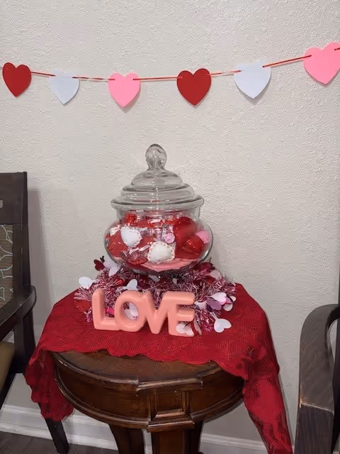A small wooden table covered with a red cloth, holding a glass jar filled with red, white, and pink decorative balls. In front of the jar is a pink decorative sign spelling the word 'LOVE'. Above the table, a string of red, white, and pink heart-shaped cutouts is hung on the wall. Two chairs are partially visible on either side of the table.