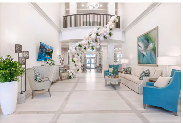 Bright and spacious lobby area with high ceilings and white walls, decorated with a large balloon garland hanging from the upper balcony. The seating area includes a white sofa, blue armchairs, and a coffee table. There are large abstract paintings on the walls and potted plants adding greenery to the space.
