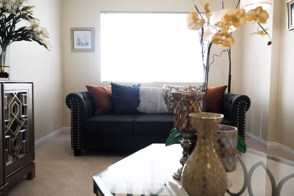 A cozy living room with a dark sofa adorned with multiple cushions in various colors and patterns. In front of the sofa is a glass coffee table with decorative vases and flowers. To the left, there is a cabinet with a vase of white flowers on top. A large window behind the sofa lets in bright natural light, and the walls are decorated with framed artwork.