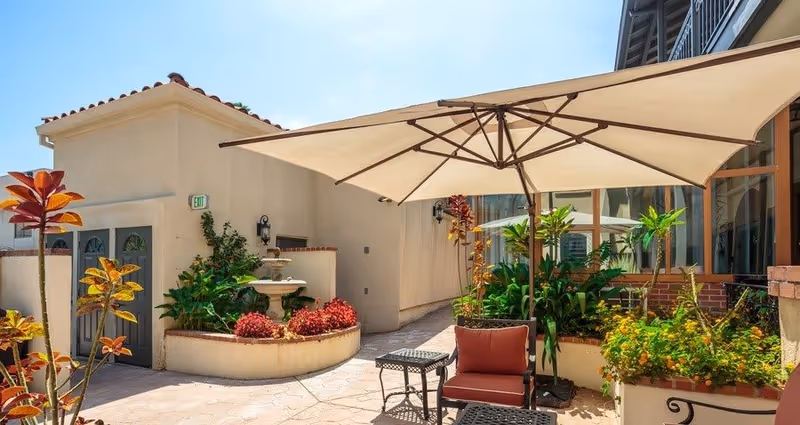 Outdoor patio area with a large beige umbrella shading a red cushioned chair and a small black metal table. Surrounding the patio are various plants and flowers in planters, with a beige building wall and windows in the background. The sky is clear and blue.
