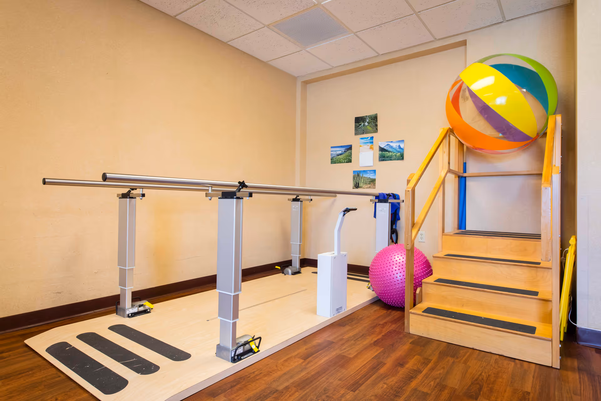 Physical therapy room with parallel bars for walking exercises, a set of wooden stairs with handrails, a large colorful beach ball, and a pink exercise ball. Several small landscape photos are displayed on the wall.