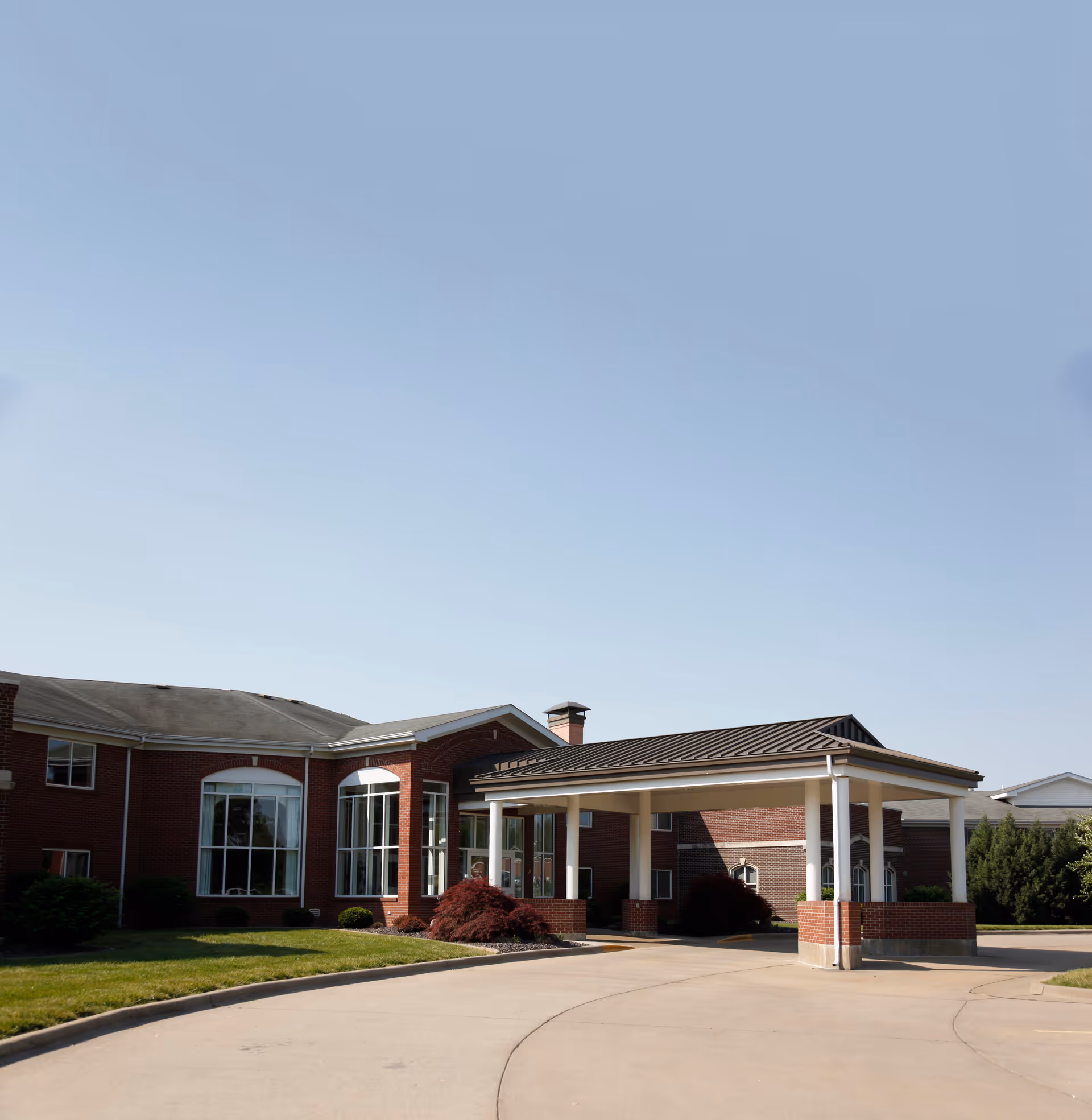 Exterior view of Saxony Village Retirement Community showing a brick building with large windows and a covered driveway entrance. The sky is clear and the surrounding area has green grass and shrubs.