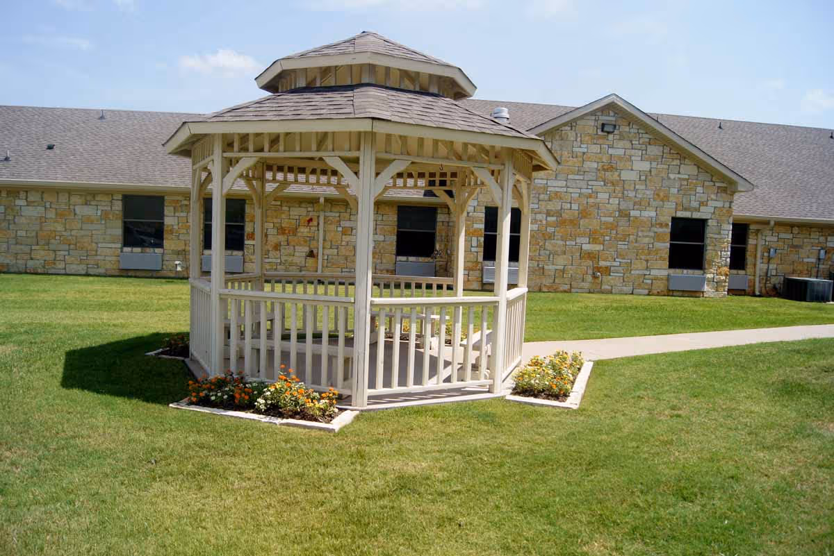 A white wooden gazebo with a shingled roof situated on a well-maintained grassy lawn with small flower beds around its base. In the background, there is a stone building with several windows and a paved walkway leading past the gazebo.