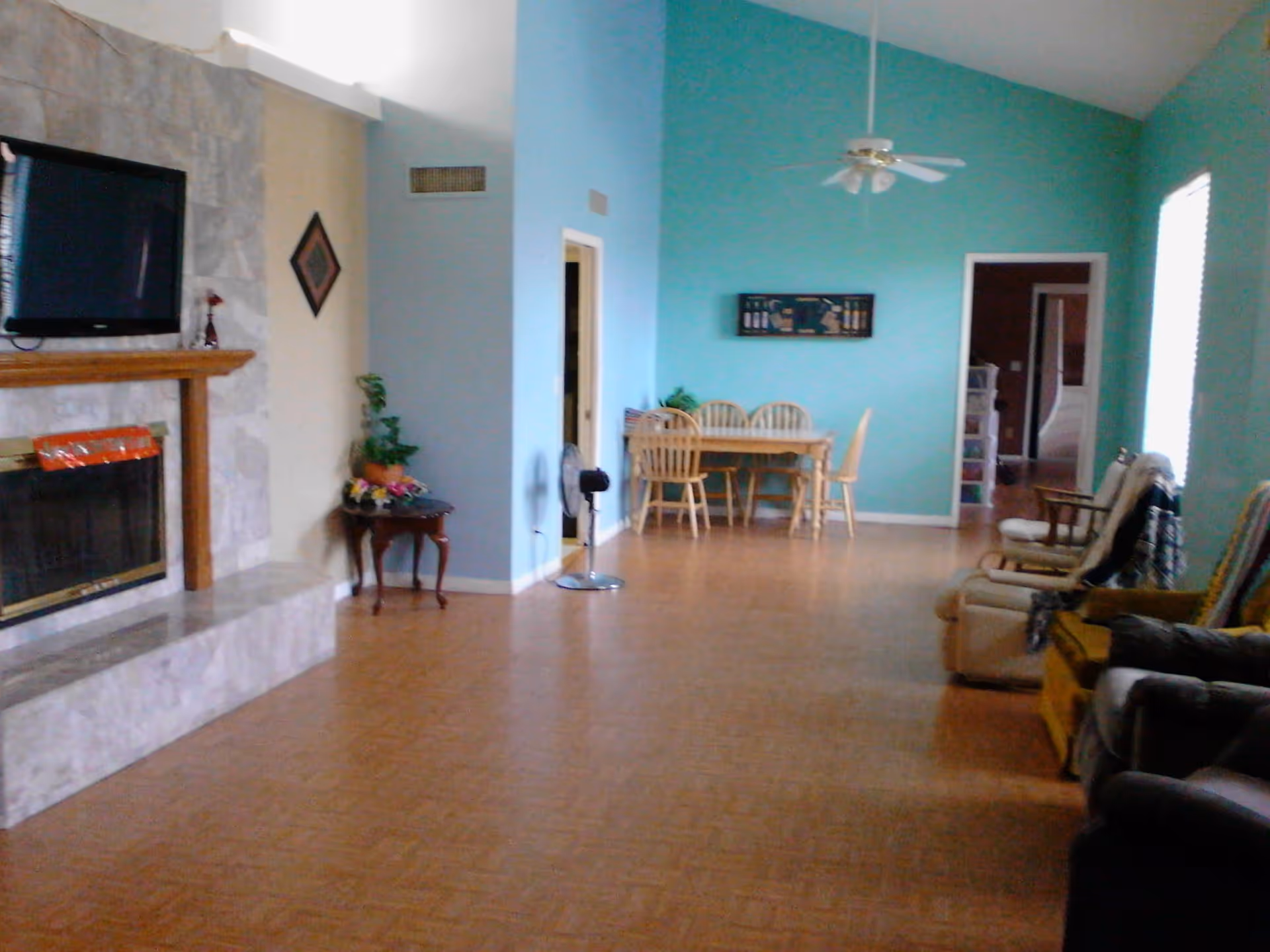 Open living room with a stone fireplace and wall-mounted TV on the left, seating on the right, and a dining table against a teal wall in the background.