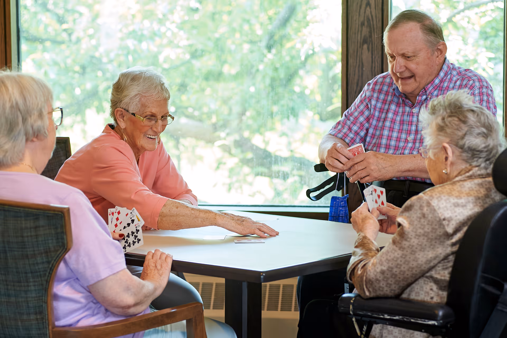 Four elderly people sitting around a table playing cards in a well-lit room with large windows showing green trees outside. They appear engaged and happy, with one woman reaching out to place a card on the table.