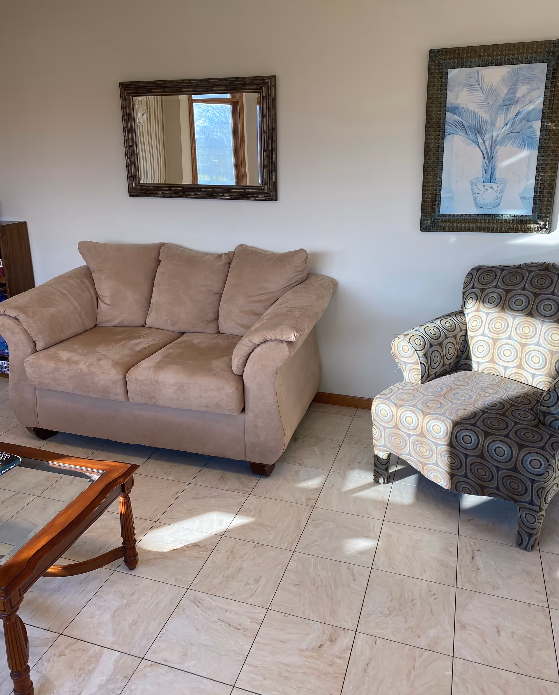 A cozy living room area with a beige loveseat and a patterned armchair on a tiled floor. A wooden coffee table with a glass top is partially visible in the foreground. On the wall above the loveseat, there is a rectangular mirror reflecting a window, and to the right, a framed artwork of a blue-toned potted plant.