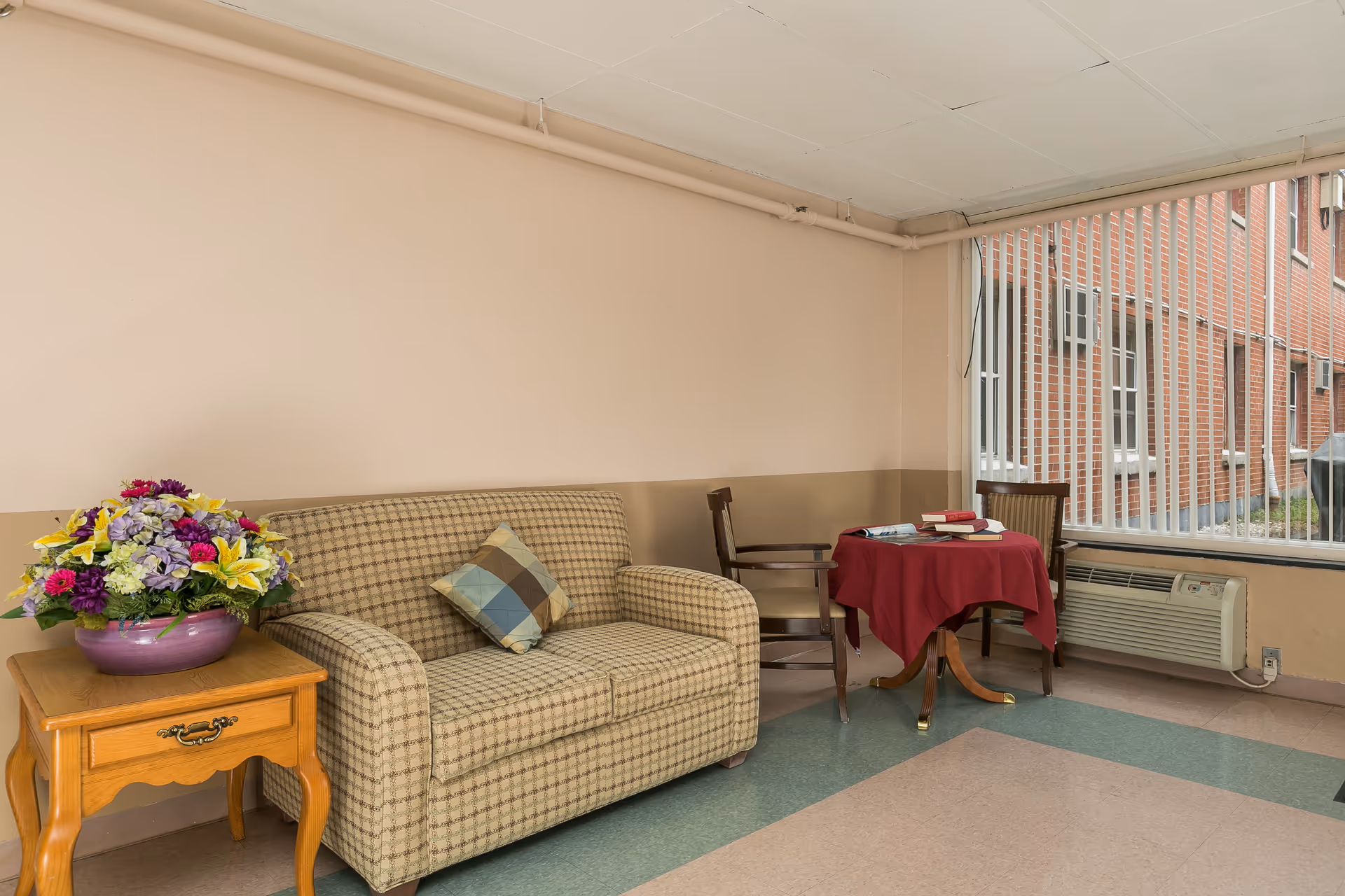 Small lounge with a patterned loveseat, side table holding a floral arrangement, and a round table with chairs by a large window with vertical blinds.