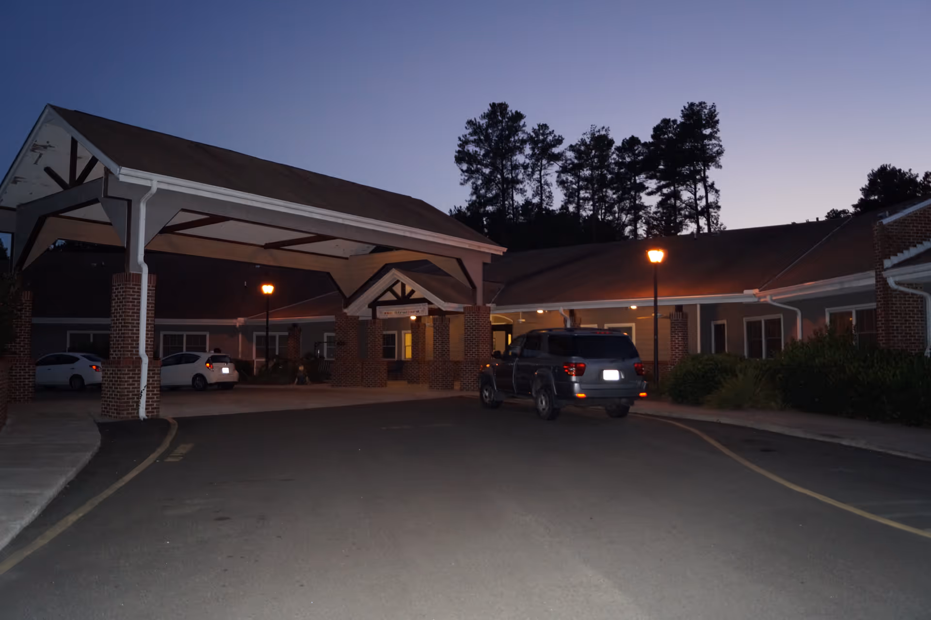 Exterior view of The Stratford senior living facility at dusk, showing a covered entrance with brick pillars, a driveway with parked cars, and trees silhouetted against the evening sky.