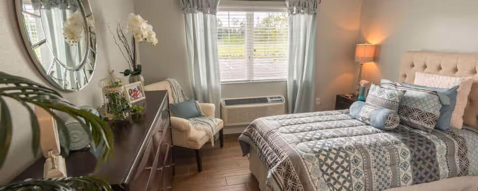 A cozy bedroom with a neatly made bed featuring patterned bedding and multiple pillows. There is a beige upholstered headboard, a nightstand with a lamp, and a window with light curtains letting in natural light. A dresser with decorative items and a round mirror is on the left side, along with a comfortable armchair near the window.