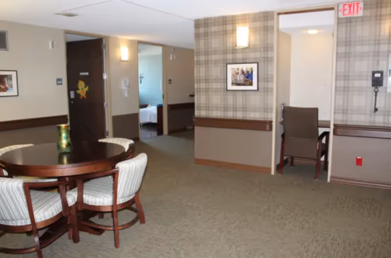 Interior view of a senior living facility common area with a round wooden table surrounded by four cushioned chairs. The walls are decorated with framed pictures and a plaid wallpaper accent. There are doorways leading to other rooms, including one with a bed visible and another with a chair and desk. An exit sign is visible above one doorway.