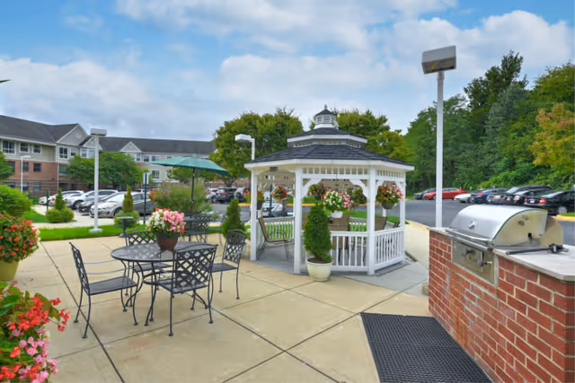 Outdoor patio area at Cascades Village featuring a white gazebo decorated with hanging flower baskets, black metal tables and chairs with flower pots, a brick barbecue grill, and a parking lot with cars in the background under a partly cloudy sky.