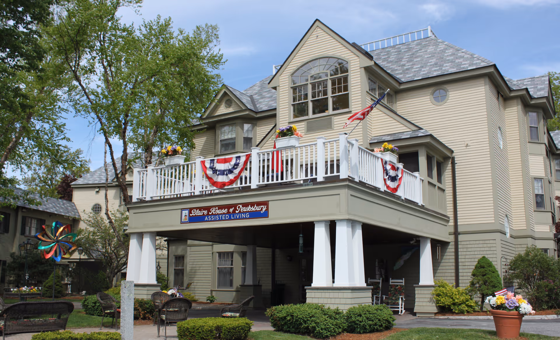 Exterior view of Blaire House of Tewksbury assisted living facility, showing a large beige building with a balcony decorated with American flag bunting and flower pots. There are trees, bushes, outdoor chairs, and a colorful pinwheel in the foreground under a blue sky.