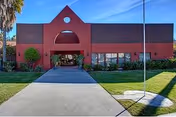 Red-brick building facade with a central arched entrance, driveway, and landscaped lawn in front.