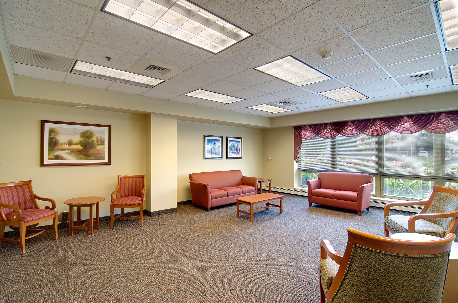 A spacious senior living facility common area with beige walls and carpeted floor. The room features two red sofas, several wooden chairs with cushions, small wooden tables, and large windows with purple valance curtains letting in natural light. Three framed landscape paintings hang on the walls.