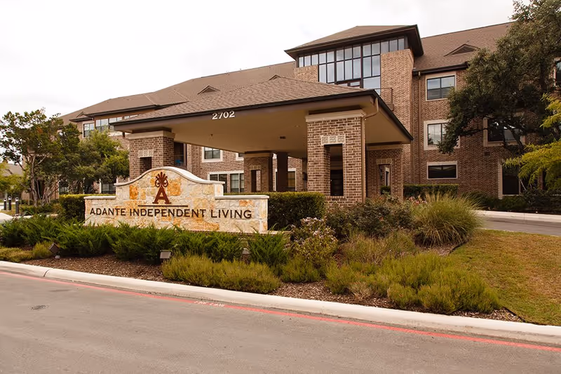 Exterior view of Adante Independent Living facility showing a brick building with multiple windows, a covered entrance with pillars, and a stone sign with the facility's name surrounded by landscaped greenery.
