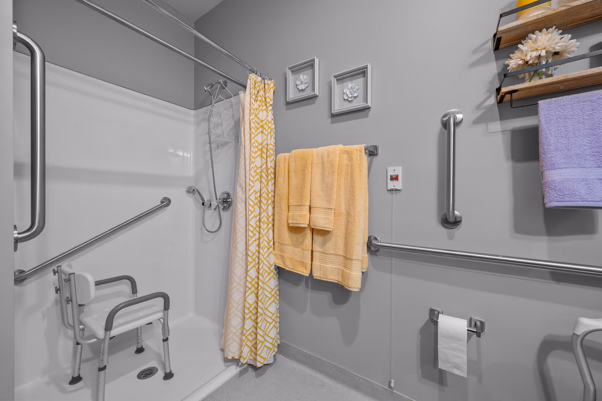 Accessible bathroom featuring a roll-in shower with a shower chair, multiple grab bars, and yellow towels hanging on the wall.