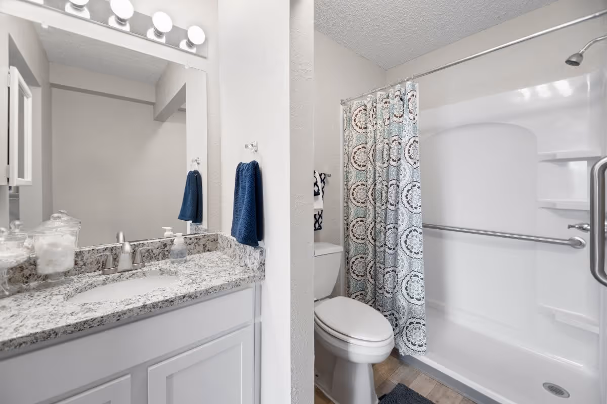 A clean and modern bathroom featuring a granite countertop with a sink, a large mirror with four round lights above it, a toilet, and a bathtub with a patterned shower curtain. The bathtub has a grab bar installed for safety.