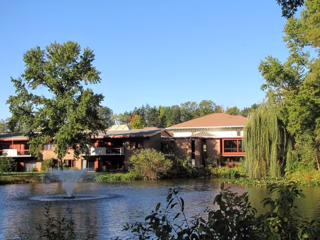 View of a senior living facility building named Meadow Lakes, situated behind a pond with a water fountain. The scene includes large trees, greenery, and a clear blue sky.