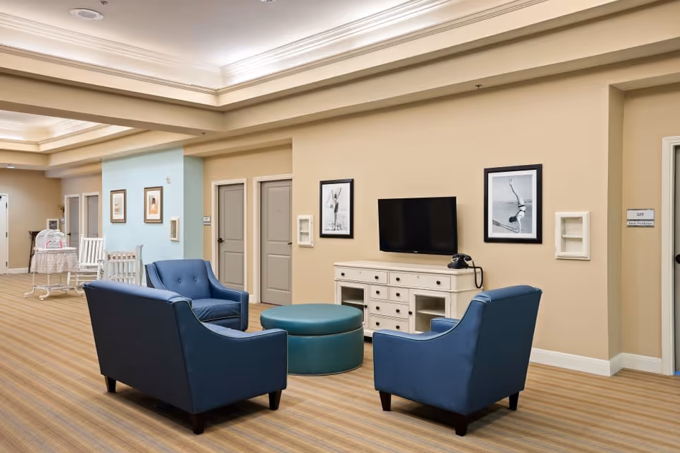 Interior common lounge with three blue armchairs and a round ottoman facing a wall-mounted TV and console.