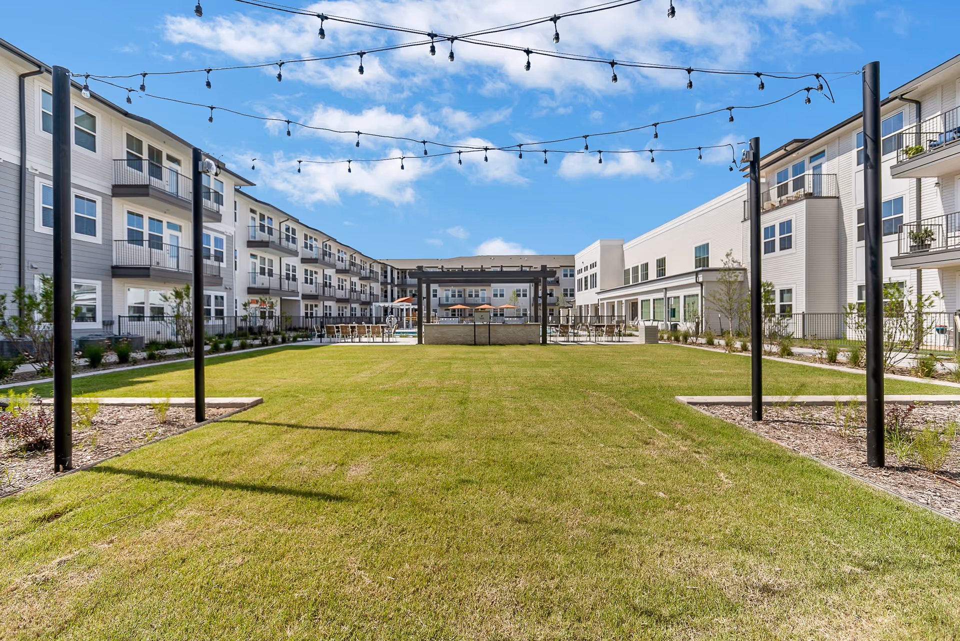 A central grassy courtyard with hanging string lights surrounded by three-story senior living buildings and outdoor seating.