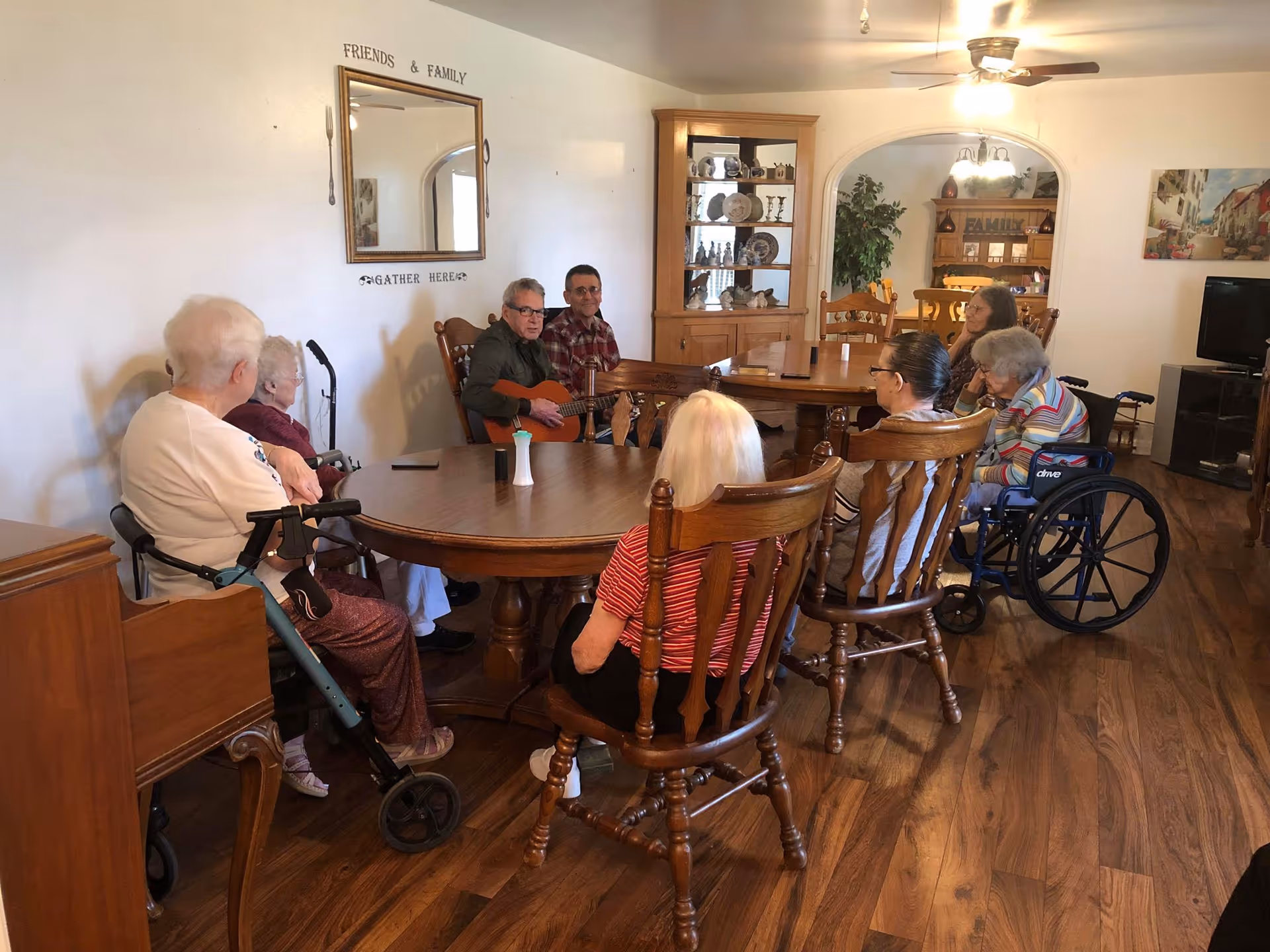 A group of elderly people sitting around a large wooden table in a living room or common area. Two men are playing guitars while others listen attentively. The room has wooden flooring, a wooden cabinet with decorative plates, a mirror on the wall with the words 'FRIENDS & FAMILY GATHER HERE', and a television in the background.