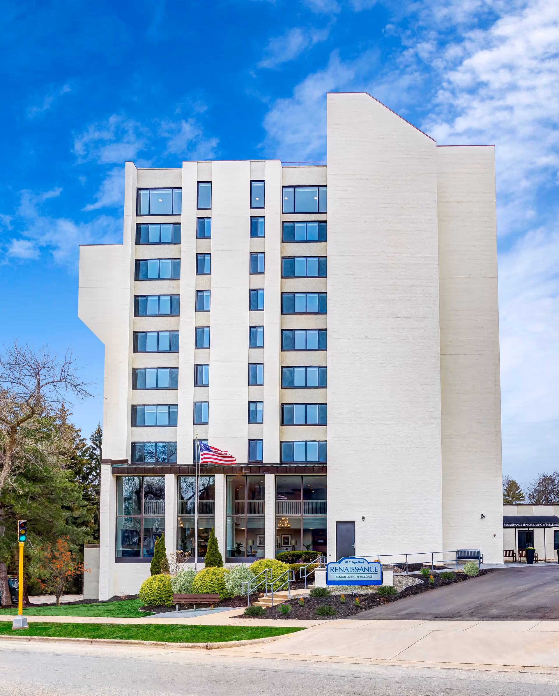 Tall white multi-story retirement community building with many windows, an American flag, entry landscaping and a sign reading Renaissance in front under a blue sky.