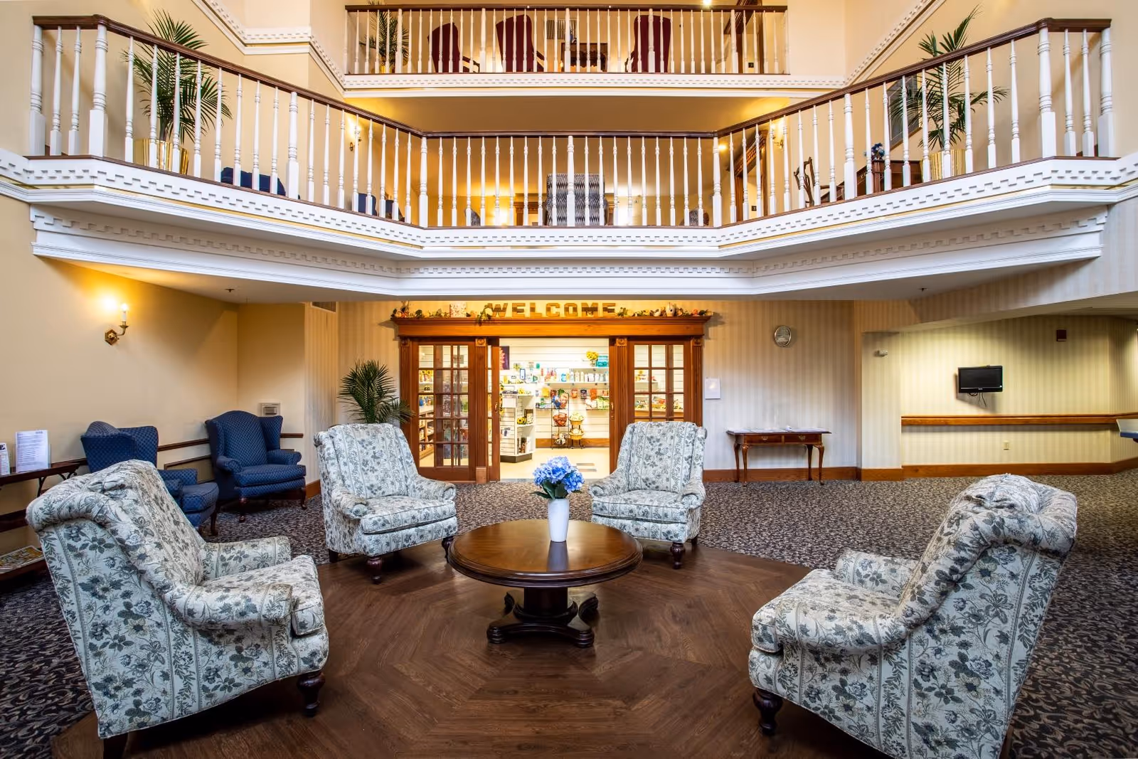 A spacious senior living community lounge area with four floral upholstered armchairs arranged around a round wooden coffee table with a vase of blue flowers. The room features a high ceiling with white railings on the upper floors, warm lighting, and a carpeted floor with a wooden section under the seating area. In the background, there is a wooden-framed glass door entrance with a 'WELCOME' sign above it and a glimpse into a well-lit room beyond.
