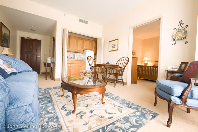 Furnished living area with a blue sofa, wooden coffee table, small kitchenette and a dining set, with a bedroom visible through a doorway.