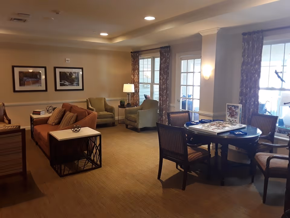 A cozy living room area in a senior living facility with a brown sofa, two green armchairs, a round table with four chairs, framed pictures on the wall, and large windows with patterned curtains letting in natural light.