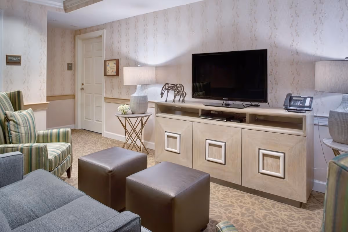 Cozy living room with a TV on a wooden console, striped armchairs, ottomans, side tables and lamps in a senior facility.