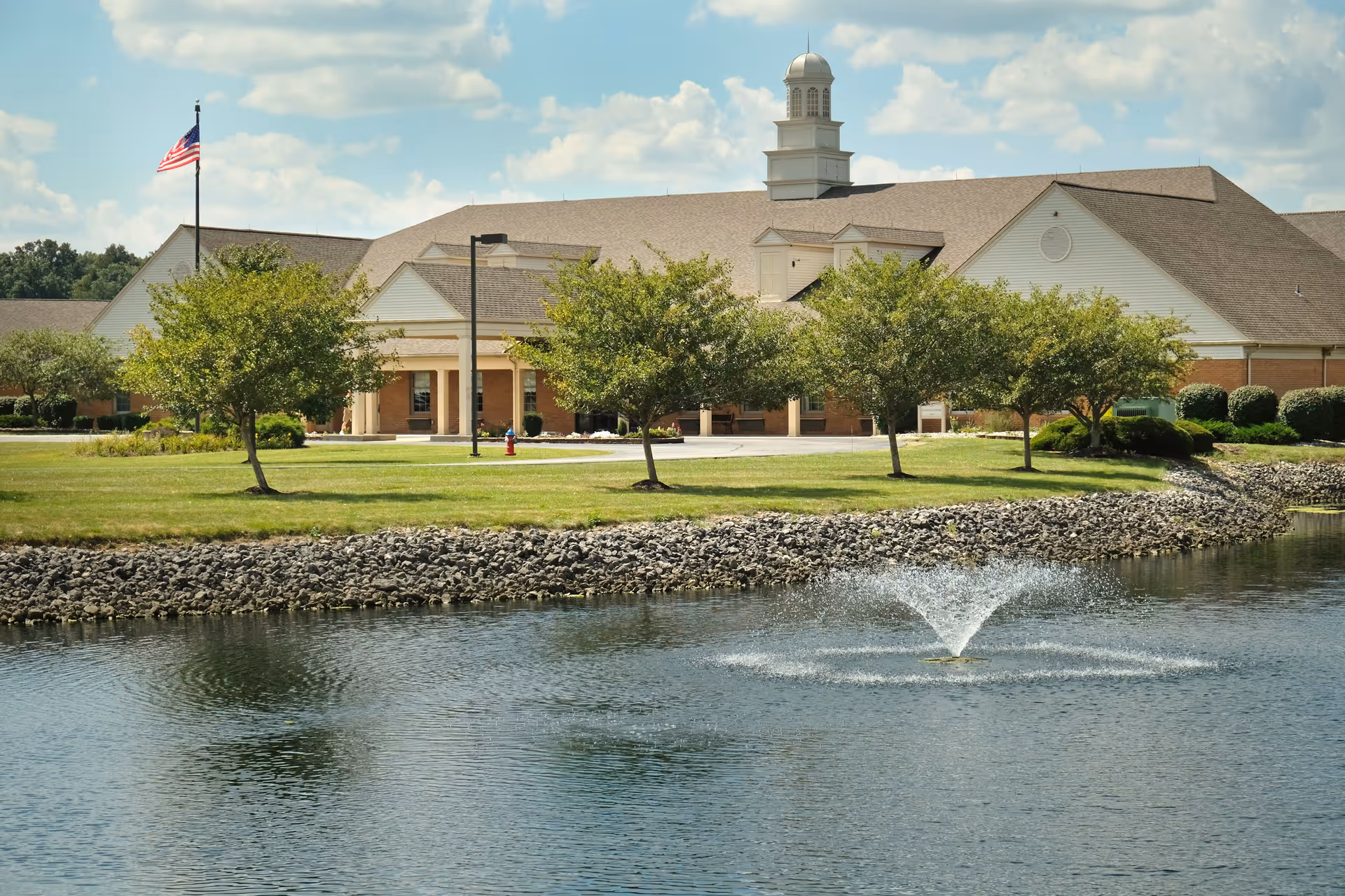 Exterior view of Otterbein Cridersville SeniorLife Community building with a pond and water fountain in the foreground, green lawn, several trees, and an American flag on a flagpole under a partly cloudy sky.