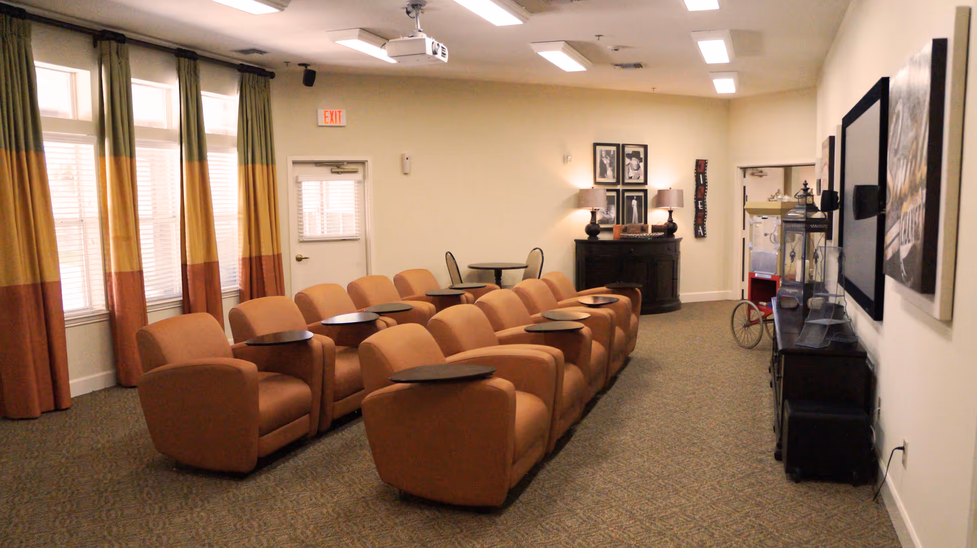 A cozy common area with two rows of orange armchairs equipped with small attached tables, facing a wall-mounted TV. The room has large windows with colorful curtains in green, yellow, and orange, a carpeted floor, and soft lighting from lamps on a side cabinet. There is also a popcorn machine and framed pictures on the walls.