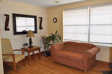 Cozy living room area with sofa, chair, lamp, plants, and large windows.
