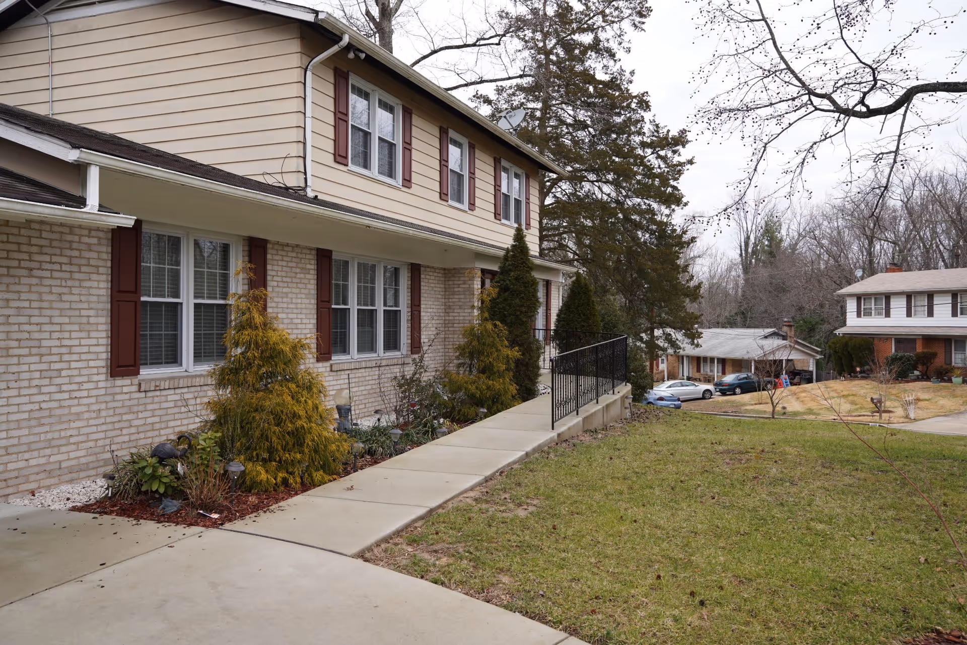 Exterior view of a two-story residential building with beige siding and brick walls, red shutters on the windows, a concrete walkway with a black metal railing, and landscaped bushes along the side. In the background, there are other houses, trees without leaves, and a cloudy sky.