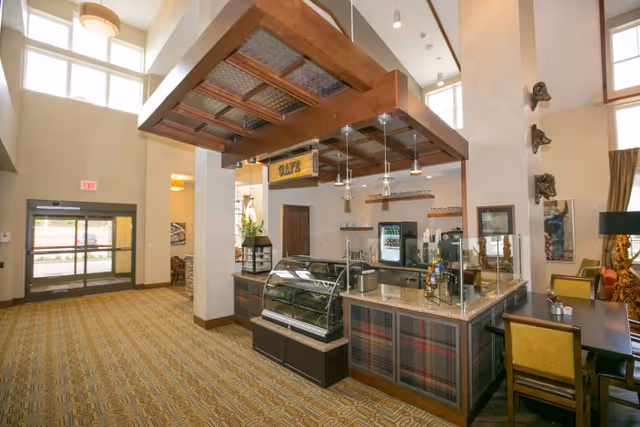 Interior view of a cafe area inside a senior living facility with a display case, counter, and seating area. The space features high ceilings, pendant lighting, and a patterned carpet. There is a glass door entrance visible in the background.