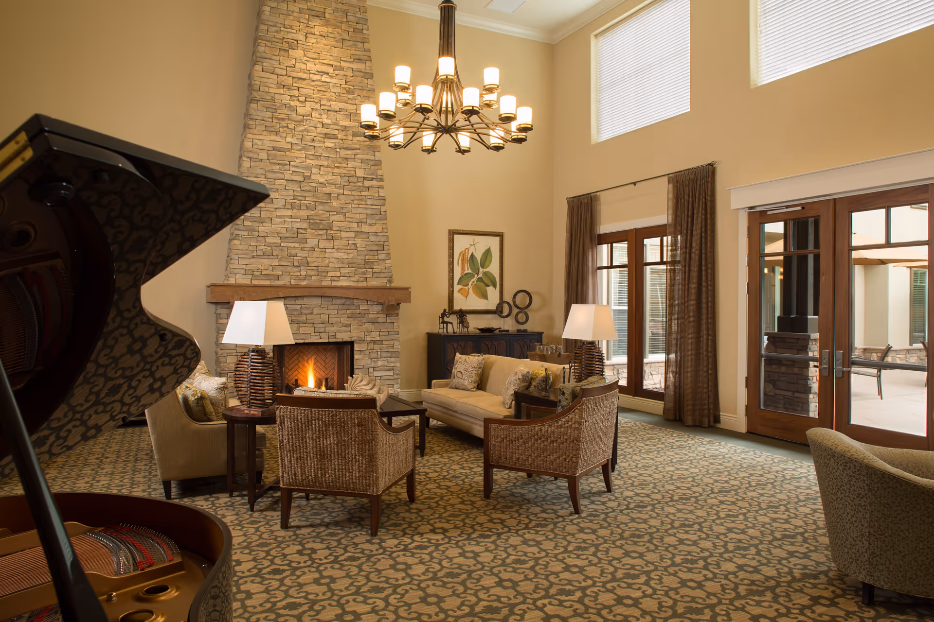 A spacious living room with a high ceiling, featuring a stone fireplace with a fire burning, a chandelier, a grand piano, a sofa, armchairs, side tables with lamps, a framed botanical artwork, and large windows with brown curtains overlooking an outdoor patio.