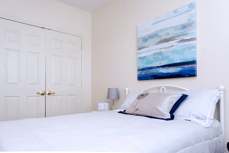 A neatly made bed with white bedding and a decorative pillow featuring a flower design in a bedroom. Above the bed hangs a large abstract painting with blue and white tones. To the left of the bed is a small nightstand with a gray lamp and a framed picture. In the background, there are double white closet doors with gold handles.