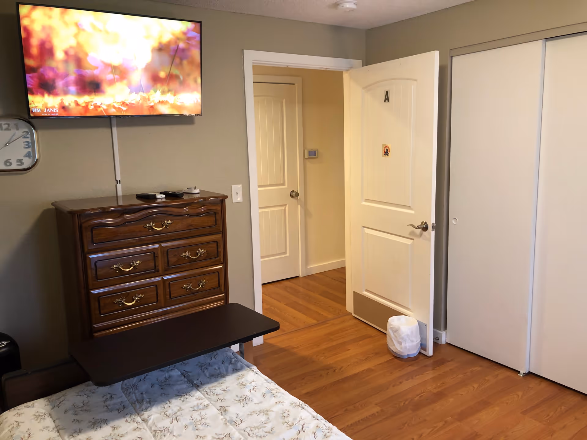 A bedroom with a wooden dresser, a wall-mounted TV displaying a colorful autumn scene, a clock on the wall, a bed with a floral bedspread, a small table over the bed, and an open door labeled 'A' leading to a hallway. There is a trash can near the door and a large white closet with sliding doors.
