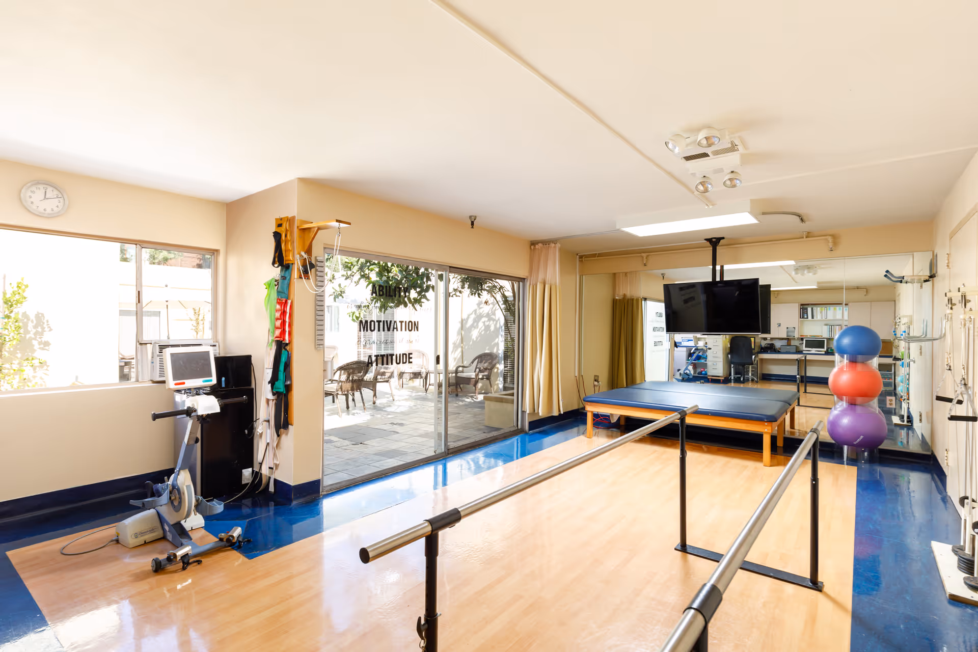 A bright physical therapy room with wooden flooring and blue borders. The room features parallel bars for walking exercises, a stationary exercise bike, a large padded therapy table, a wall-mounted TV, and colorful exercise balls stacked in a corner. Large windows and sliding glass doors provide natural light and a view of an outdoor patio with chairs. The walls have motivational words like 'ABILITY', 'MOTIVATION', and 'ATTITUDE'.