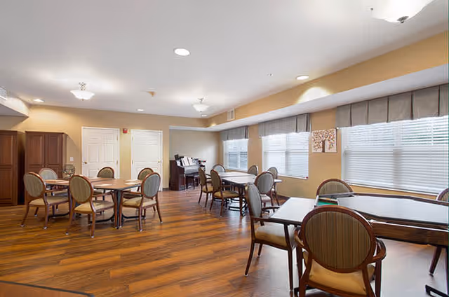 A well-lit dining room with several wooden tables and cushioned chairs arranged neatly. The room has large windows with blinds and valances, wooden flooring, and a piano in the corner. The walls are painted in a warm beige tone.