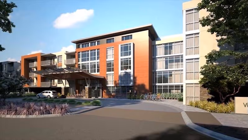 Exterior view of a modern multi-story senior living facility with large windows, a covered entrance, parked cars, and bicycles near the entrance. The building is surrounded by landscaping with trees and shrubs under a clear blue sky.