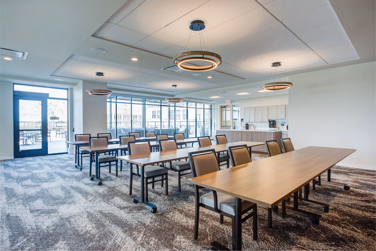 A bright and spacious dining room with multiple wooden tables and cushioned chairs arranged neatly. The room features large windows allowing natural light to fill the space, modern circular ceiling lights, and a door leading to an outdoor patio area with additional seating. There is also a counter area with cabinets and a coffee machine in the background.