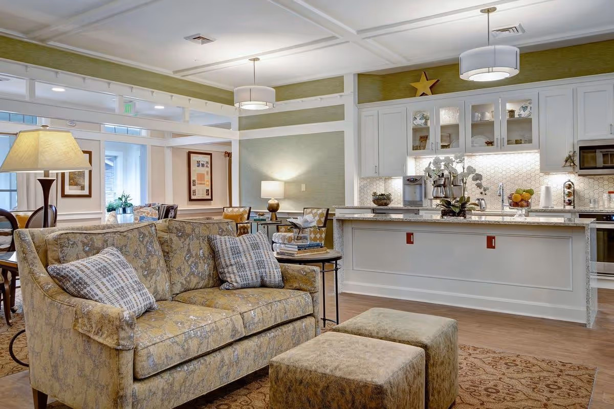 Bright open communal living area with a patterned sofa and ottomans facing a kitchen island with white cabinetry and pendant lights.