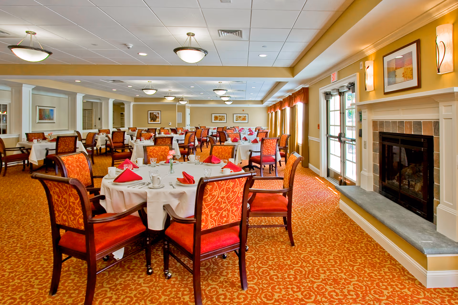 Bright carpeted dining room with round tables set with white linens and red napkins, upholstered chairs, and a fireplace near glass doors.