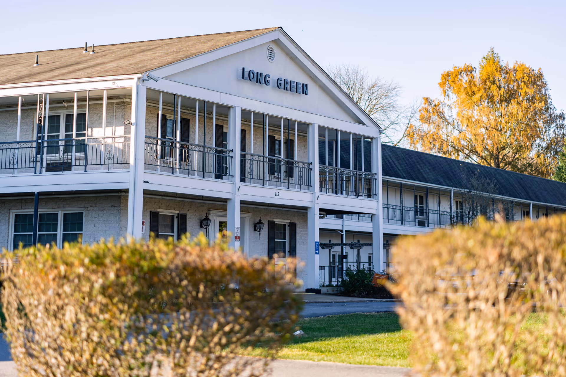 Two-story white senior living building with balconies and a "LONG GREEN" sign, seen behind trimmed hedges.